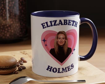 May include: A white ceramic mug with a dark blue interior and handle. The front features a pink heart graphic containing a photograph of Elizabeth Holmes, surrounded by small star icons. The text ELIZABETH is printed above and HOLMES below the heart.