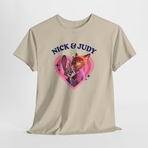 K&ouml;nnte beinhalten: Beiges T-Shirt mit einem Aufdruck von Nick Wilde und Judy Hopps aus Zootopia in einem rosa Herz. Der Text "NICK & JUDY" ist blau. Das Design umfasst rosa und wei&szlig;e konzentrische Kreise und blaue Sternakzente.