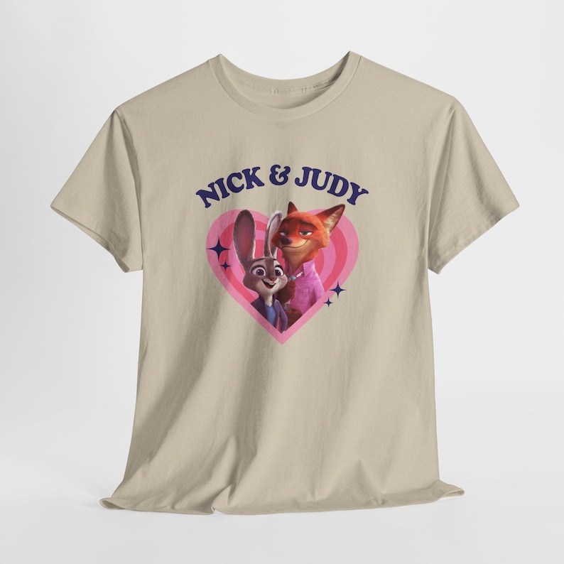 K&ouml;nnte beinhalten: Beiges T-Shirt mit einem Aufdruck von Nick Wilde und Judy Hopps aus Zootopia in einem rosa Herz. Der Text "NICK & JUDY" ist blau. Das Design umfasst rosa und wei&szlig;e konzentrische Kreise und blaue Sternakzente.
