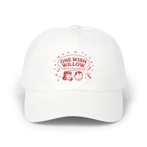 May include: White baseball cap with a curved brim. The cap features a red graphic with the words "ONE WISH WILLOW" and the phrase "AMAZE YOUR FRIENDS". The graphic includes illustrations of a woman and a man.