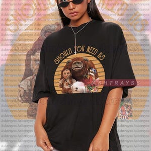 Op de afbeelding: Zwart unisex T-shirt met een afbeelding uit de film "Labyrinth" en de tekst "SHOULD YOU NEED US". Het ontwerp bevat personages en een cirkelvormig logo. Het model draagt een 2XL shirt.