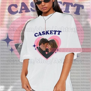 May include: White unisex t-shirt with the word "CASKETT" in purple, and a heart graphic with a couple embracing. The shirt is oversized. The model is wearing a 2XL.