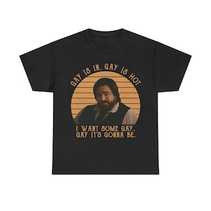 May include: A black short-sleeved t-shirt featuring a circular graphic with a sunset-style orange striped background. The graphic shows a bearded person in a vest and white shirt, with the text GAY IS IN. GAY IS HOT. I WANT SOME GAY. GAY IT'S GONNA BE.