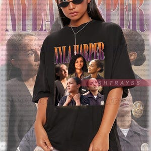May include: A black t-shirt with a vintage-style graphic of a group of people, including a woman with a cross necklace, and the text "NYLA HARPER" in a bold, colorful font. The shirt is oversized and features a faded, distressed look.