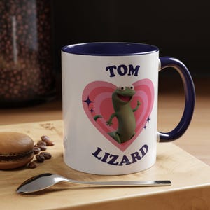 Könnte beinhalten: Weiße Keramiktasse mit marineblauem Innenraum und Griff. Die Tasse zeigt eine Cartoon-Eidechse in einem rosa Herzen mit den Worten "TOM LIZARD" in Blau. Ein Löffel und ein Macaron befinden sich im Vordergrund.