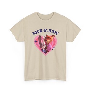 K&ouml;nnte beinhalten: Beiges T-Shirt mit dem Text "NICK & JUDY" &uuml;ber einem rosa Herz. Das Herz enth&auml;lt Cartoon-Bilder eines Kaninchens in einer blauen Weste und eines Fuchses in einem rosa Hemd.
