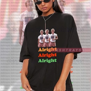 May include: Black unisex t-shirt featuring a graphic of three people with the text "Alright Alright Alright" in orange, red, and green. The shirt is oversized and has a crew neck.