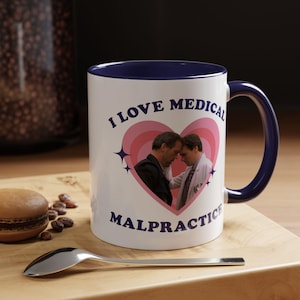 Puede incluir: Taza de cerámica blanca con borde y asa azules. La taza presenta un corazón rosa con dos hombres dentro y el texto "Me encanta la mala praxis médica".
