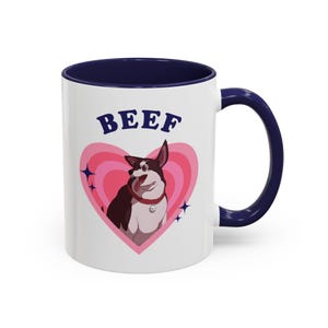 Puede incluir: Taza de cerámica blanca con interior y asa azul marino. La taza presenta un perro de dibujos animados dentro de un corazón rosa con la palabra "BEEF" encima. El perro es marrón y blanco con un collar rojo.