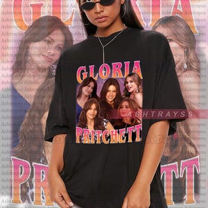 Puede incluir: Camiseta negra unisex con el texto "GLORIA PRITCHETT" y un collage de rostros de mujeres. La camiseta es un homenaje al personaje de la serie de televisión. La modelo lleva 2XL.