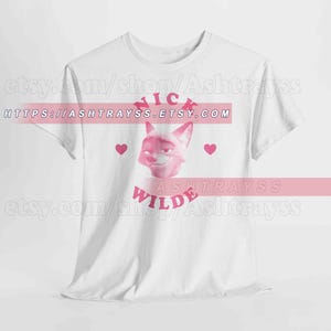 May include: White t-shirt featuring a pink illustration of a fox's face with the name "Nick Wilde" in pink text. The design includes pink hearts. The Etsy shop URL is visible.