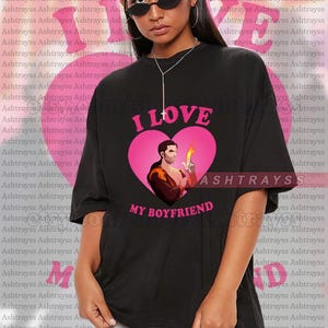 May include: Black t-shirt with a pink heart graphic that says "I LOVE MY BOYFRIEND". The graphic features an illustration of a man holding a flame. The shirt is a unisex style.