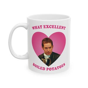 Puede incluir: Taza de cerámica blanca con asa. Presenta un corazón rosa con el retrato de un hombre y el texto "WHAT EXCELLENT BOILED POTATOES" en rosa. Taza de tamaño estándar para café o té.