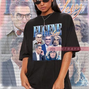 May include: Black unisex t-shirt featuring a collage of Eugene Levy in various roles, with the text "EUGENE LEVY" in a blue and white design. The shirt is part of a collection of apparel.