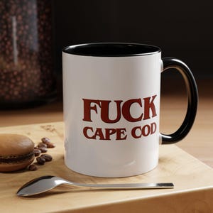 Pode incluir: Uma caneca de cerâmica branca com interior e alça pretos, apresentando o texto em vermelho escuro "FUCK CAPE COD" impresso na frente. A caneca é cilíndrica com um acabamento liso e uma alça em forma de C, exibindo letras claras e ousadas.