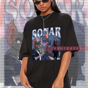 May include: Black oversized t-shirt with a graphic of bat-like creatures, the word "SONAR" above and "ASHTRAYSS" below. The shirt is worn by a person with dark hair and sunglasses.