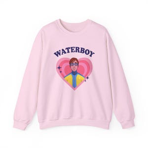Puede incluir: Sudadera rosa claro con un gráfico. El gráfico presenta la palabra "WATERBOY" encima de un diseño en forma de corazón. Dentro del corazón hay una ilustración de una persona con gafas de sol y una chaqueta amarilla y azul.
