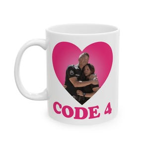 May include: White ceramic mug with a pink heart design containing a photo of a man and woman hugging. The mug has the words "CODE 4" in pink. The mug is a standard size with a white handle.