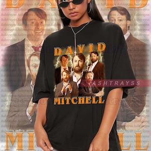 May include: Black unisex t-shirt featuring a graphic of David Mitchell and the text "DAVID MITCHELL" in orange. The shirt has a vintage style and is true to size. The model is wearing a 2XL.
