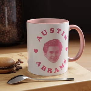 May include: A white ceramic mug with a pink interior and handle. The front features a pink graphic of a person's face centred between the text AUSTIN in an arc above and ABRAMS in an arc below, with small pink hearts on the left and right sides.