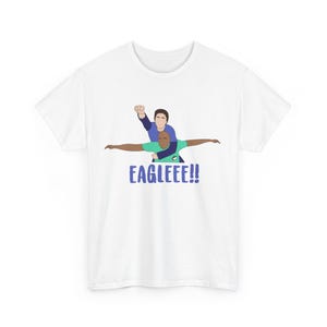 May include: White t-shirt featuring a cartoon illustration of two people in a celebratory pose, with one person on top and the other with arms outstretched. The text "EAGLEEE!!" is printed in blue below the figures.