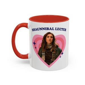 May include: White ceramic mug with a red interior and handle. The mug features the text "SHAUNNIBAL LECTER" above an image of a woman inside a pink heart design. The design includes blue star accents.