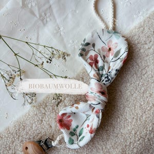 Puede incluir: Pinza para chupete floral con una cuenta de madera y un clip metálico. La tela es blanca con un estampado de flores rosas, rojas y verdes. El texto "BIOBAUMWOLLE" es visible.