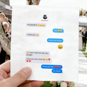 Könnte beinhalten: Eine weiße Papierserviette mit einer gedruckten SMS-Konversation. Die Nachrichten enthalten Sätze wie "Hallo Schönheit", "Ich vermisse dich" und "Ich liebe dich". Der Hintergrund zeigt eine Hochzeitszeremonie.