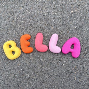 Könnte beinhalten: Fünf einzelne Buchstaben, die den Namen BELLA bilden, jeweils in einer anderen Farbe und mit einer subtilen topografischen Linienstruktur. Die Buchstaben sind gelb, orange, koralle, hellrosa und magenta und liegen in einer Reihe auf grauem Grund.