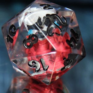 May include: A clear, multi-sided die with black numbers and a red interior design. The die is reflected in a mirrored surface, creating a symmetrical image. The background is a blurred blue.