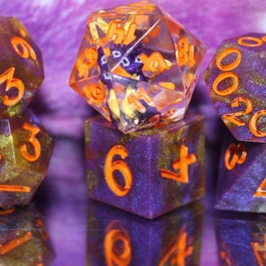May include: A collection of polyhedral dice in various shapes. The dice are purple with orange numbering and gold accents. The dice are displayed on a reflective surface, creating mirrored images.