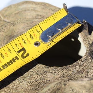 May include: A yellow HART measuring tape is positioned over a beige, textured object. The tape shows measurements in inches, with the 2-inch mark visible. The object has a natural, organic appearance, with visible wood grain patterns.
