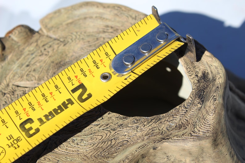 May include: A yellow HART measuring tape is positioned over a beige, textured object. The tape shows measurements in inches, with the 2-inch mark visible. The object has a natural, organic appearance, with visible wood grain patterns.