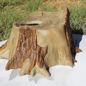 May include: A decorative tree stump-shaped container. The container is made of a light tan material with darker brown accents to mimic the look of tree bark. The top has a circular opening. The container is on a white surface.