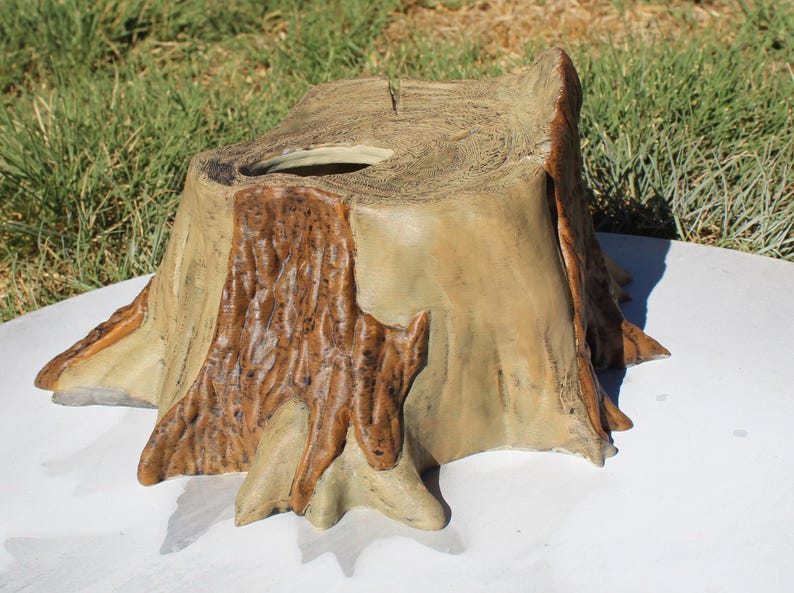 May include: A decorative tree stump-shaped container. The container is made of a light tan material with darker brown accents to mimic the look of tree bark. The top has a circular opening. The container is on a white surface.