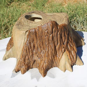 May include: A decorative, handcrafted wooden stump with a natural, textured appearance. The stump features a central hole and is painted in shades of brown and tan, with visible wood grain details. The base is on a white surface.