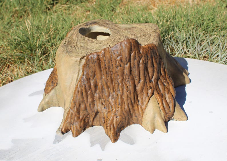May include: A decorative, handcrafted wooden stump with a natural, textured appearance. The stump features a central hole and is painted in shades of brown and tan, with visible wood grain details. The base is on a white surface.