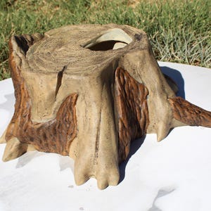 May include: A decorative tree stump planter with a hollowed-out top. The planter is painted in shades of tan and brown, with detailed wood grain and bark textures. The planter is designed to resemble a natural tree stump.