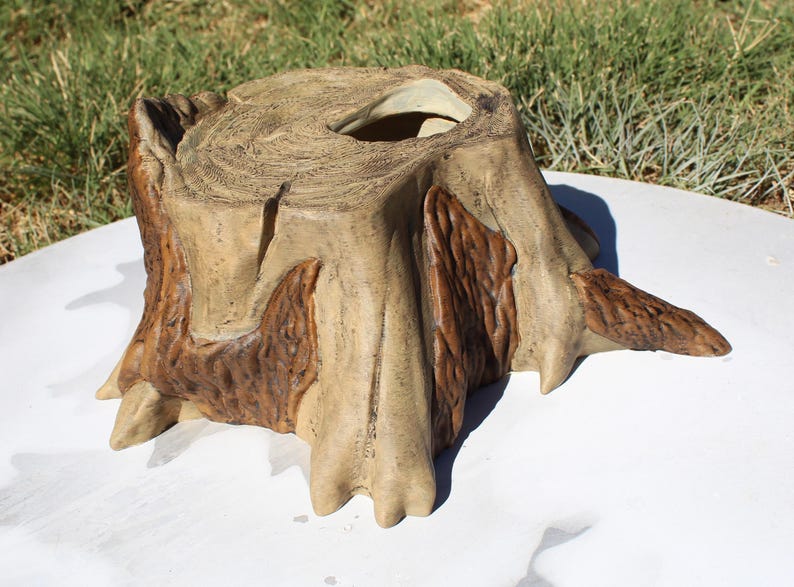 May include: A decorative tree stump planter with a hollowed-out top. The planter is painted in shades of tan and brown, with detailed wood grain and bark textures. The planter is designed to resemble a natural tree stump.