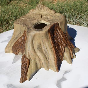 May include: A decorative tree stump-shaped container. The container is primarily tan with darker brown accents, and features a hole in the center. The design mimics the texture of tree bark and wood grain, creating a naturalistic appearance.