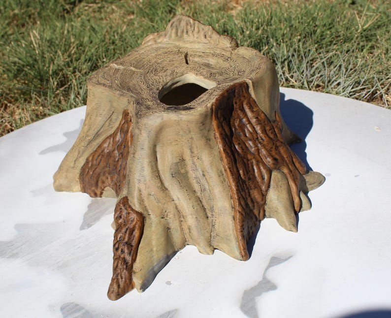 May include: A decorative tree stump-shaped container. The container is primarily tan with darker brown accents, and features a hole in the center. The design mimics the texture of tree bark and wood grain, creating a naturalistic appearance.