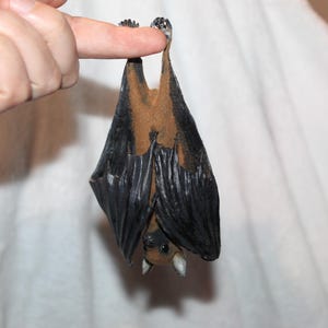 May include: A small, realistic bat figurine hanging upside down by its feet. The bat features a dark brown body with black wings that are folded against its sides. Its head is pointed downward, showing small ears and a detailed face with dark eyes.