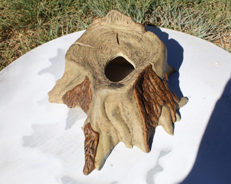 May include: A ceramic tree stump-shaped planter with a large opening. The planter is painted in shades of tan and brown, with textured details mimicking tree bark. The planter sits on a white surface with green grass in the background.