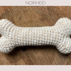 Op de afbeelding: Een gehaakt hondenbot speelgoed in een crème kleur. Het bot ligt op een houten oppervlak.