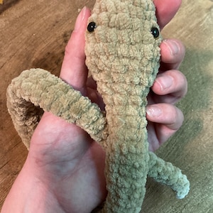 Op de afbeelding: Een gehaakt pluche speelgoed in de vorm van een worm met bruine vacht en zwarte ogen. Het speelgoed wordt in de hand van een persoon gehouden.