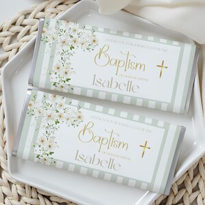 Op de afbeelding: Twee chocoladerepen met bloemmotieven en de tekst "Baptism Isabelle" worden gepresenteerd op een wit bord. De repen hebben een lichtgroene en witte gestreepte rand en gouden tekst. Het bord staat op een geweven mat.
