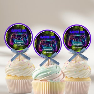 Może przedstawiać: Trzy babeczki z białym lukrem i kolorową posypką. Każda babeczka ma okrągłą nakładkę z motywem kontrolera do gier wideo oraz tekstem "GAME ON" i "ALEXANDER". Nakładki są fioletowe i zielone.