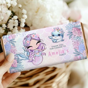Könnte beinhalten: Schokoladenverpackung mit einer Meerjungfrau und einem Delfin mit Blumenkränzen. Der Text "THANK YOU FOR COMING TO Amelia's PARTY" ist enthalten. Das Design verwendet Pastellfarben in Rosa, Blau und Lila.