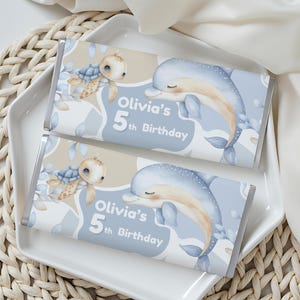 Op de afbeelding: Twee chocoladerepen met een lichtblauw en beige ontwerp. Eén reep heeft een schildpad, de andere een dolfijn. Beide repen hebben de tekst "Olivia's 5th Birthday". De repen liggen op een wit bord, bovenop een geweven mat.