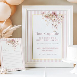 以下が含まれることがあります： 「タイムカプセル」の文字が入った、ピンクの花柄のフレーム入りサインとカード。サインにはピンクと白のストライプと金のアクセントがあります。白いケーキが台座の上にあります。